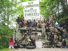 Photo shows about 20 Earth First! protesters surrounding an upturned vehicle. A large sign reads, "No Compromise on Native Forests."